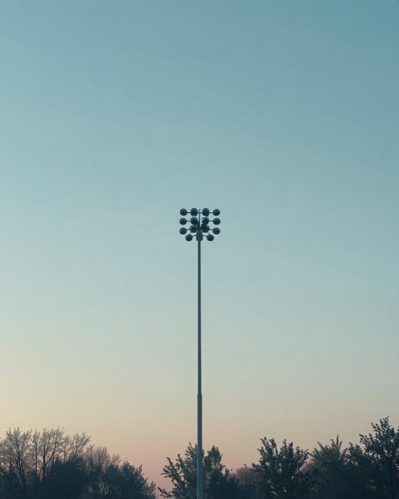 Flutlichtmast eines Sportplatzes vor hellem Abendhimmel, unten Baumkronen