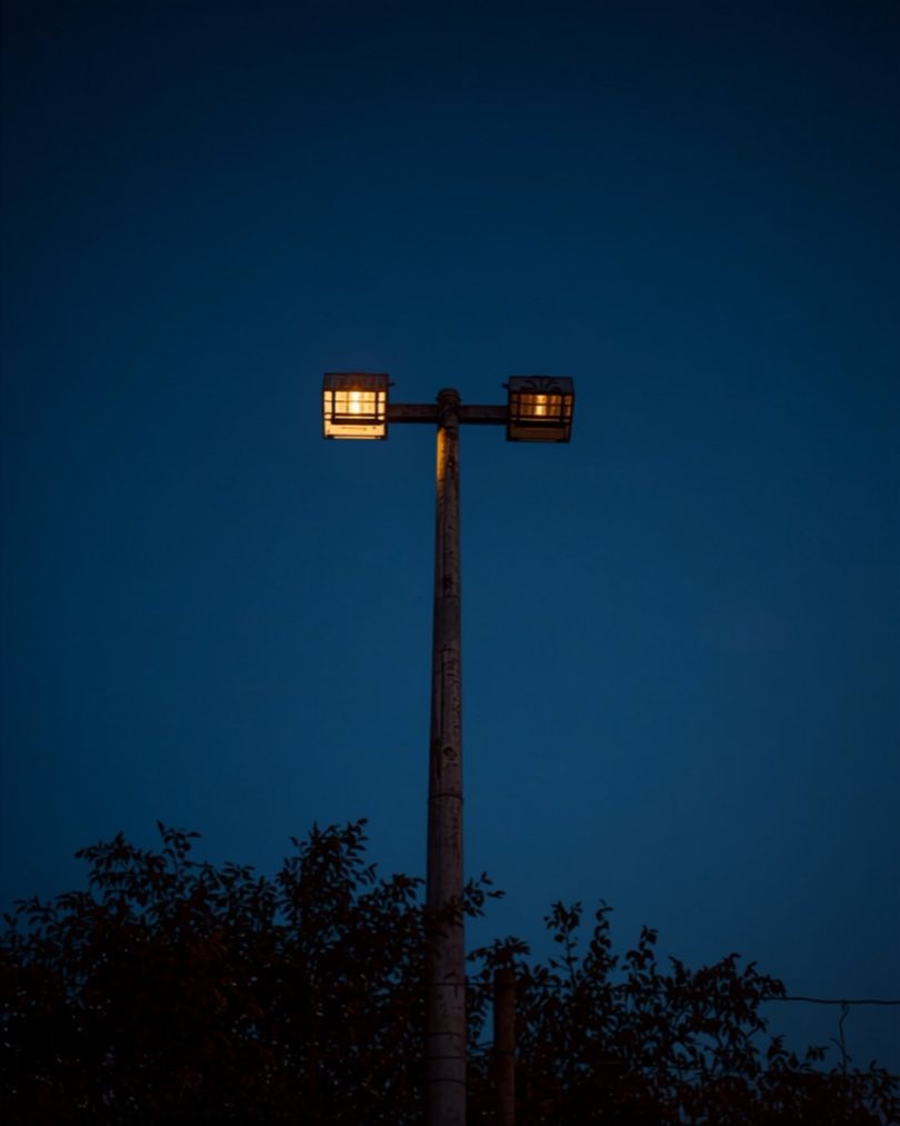 Flutlichtmast eines Sportplatzes mit zwei leuchtenden Strahlern vor dunkelblauem Abendhimmel